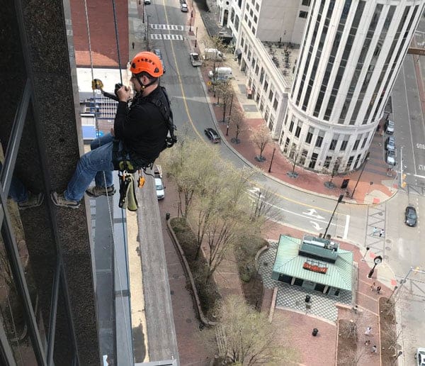 Technician in an orange helmet suspended by ropes cleaning a high-rise building with a city street view below.